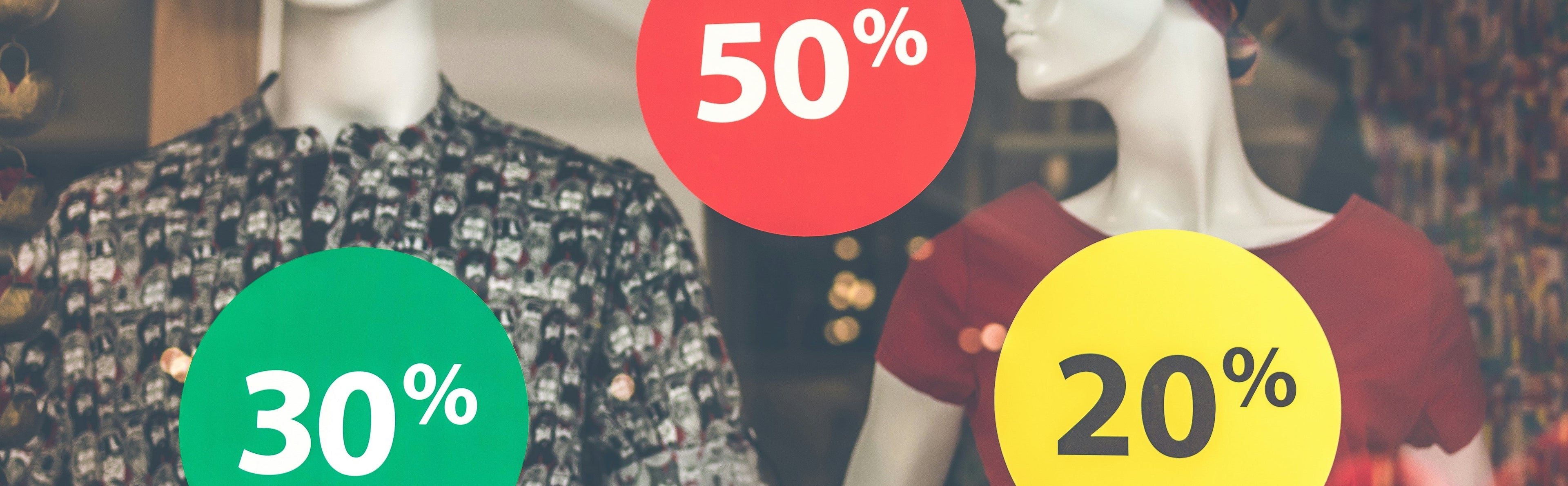 A retail window display with two mannequins wearing clothing, overlaid with large colored sale circles reading 30%, 50%, and 20% in green, red, and yellow.